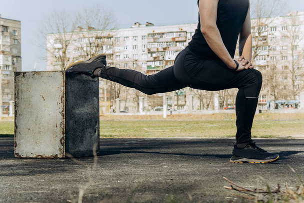 A slender young man in black clothes and sunglasses is exercising outdoors. fitness athlete on the sports field. training with projectiles. warm up stretching legs. body preparation for the summer - Foto, Bild