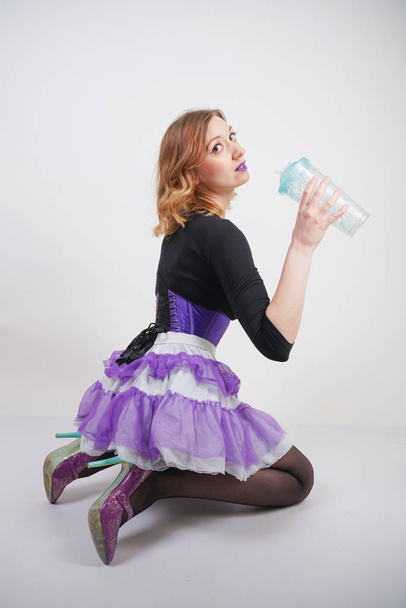 pretty slender girl in purple corset with tutu skirt and mesh black pantyhose standing with blue glass of water on white studio background - Photo, Image