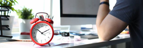 Relógio de alarme vermelho em pé na mesa de trabalho com homem pensativo
 - Foto, Imagem