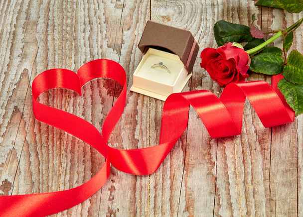 Κόκκινη κατσαρά κορδέλα σε ξύλινο φόντο με δαχτυλίδι αρραβώνων και τριαντάφυλλο - Φωτογραφία, εικόνα