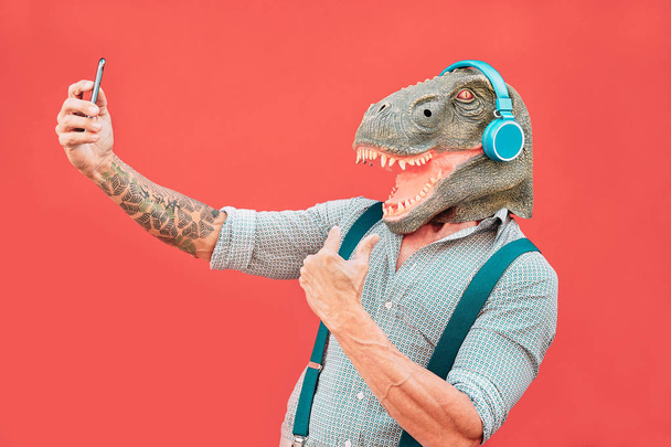 Crazy senior man wearing t-rex mask and taking selfie with mobile smartphone - Hipster older male having fun listening music and dancing outdoor -  Absurd, funny and surreal concepts - Photo, Image