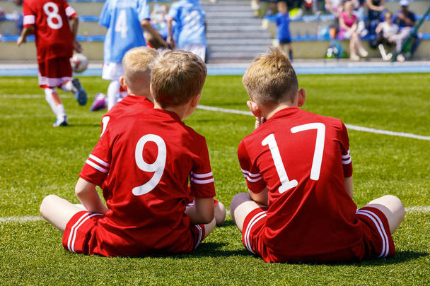 サッカーサッカーの試合をしている子供たちのグループ。サッカー場の芝生に座り、チームメイトを支える赤いサッカージャージを着た2人の若い白人少年 - 写真・画像