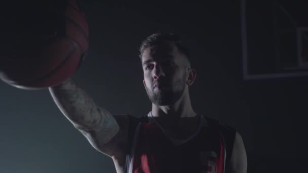 Close up portrait handsome confident man in red uniform holding a ball in outstretched hand in the dark. Professional basketball game player playing with a ball. The basket in the background - Πλάνα, βίντεο