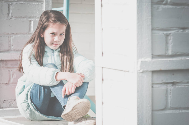 Young girl with flowing hair in blue down jacket sits and hugs her knees with her hands against background of white brick wall. - Photo, image