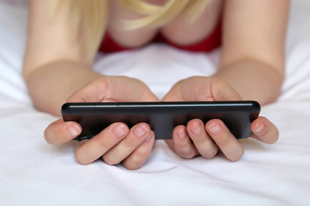 Blonde woman watching video on smartphone, sexy girl in negligee lying on the bed with mobile phone. Concept of porn, online addiction, sex dating, video chat - Valokuva, kuva
