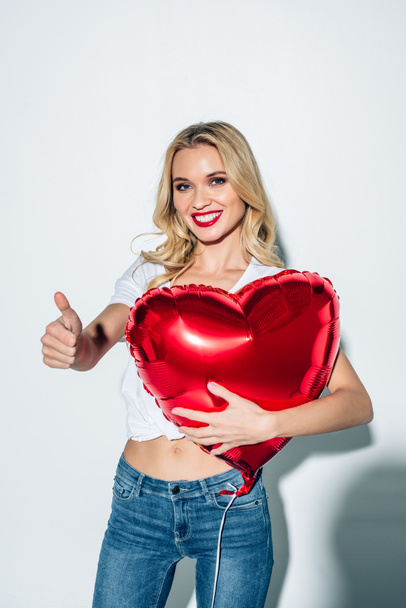 cheerful blonde girl holding red heart-shaped balloon while showing thumb up on white  - Valokuva, kuva