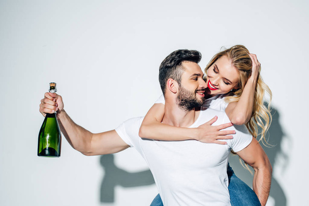 happy man holding bottle of champagne while piggybacking blonde girl on white  - 写真・画像