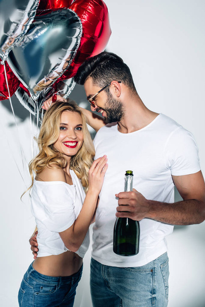 bearded man in glasses holding bottle of champagne and looking at cheerful woman with balloons on white  - Fotografie, Obrázek