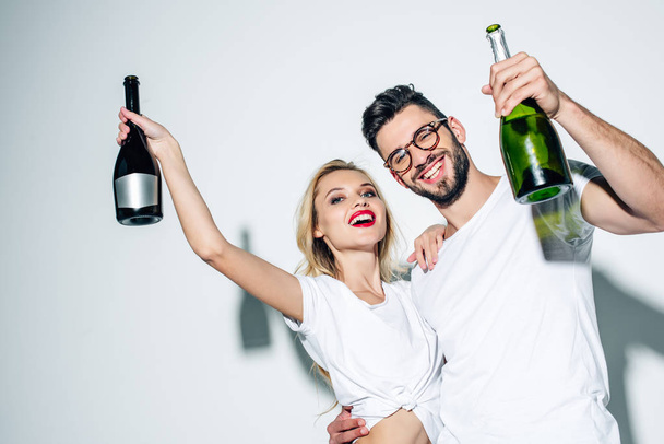 blonde girl and cheerful man in glasses holding bottles of champagne on white  - Photo, image