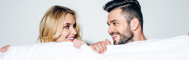 panoramic shot of happy blonde woman and cheerful man looking at each other while holding blanket on white  - Fotó, kép