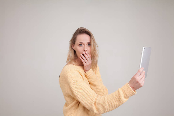 portrait of young blonde woman impressed speechless while holding her tablet in hands.wearing yellow sweater. poses against white background. face expression, emotions - Photo, Image