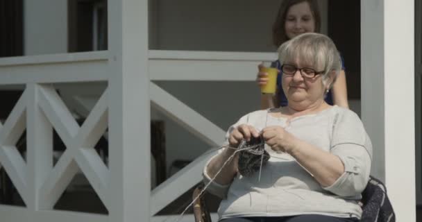 Péče o starší vnuk přináší pomerančovou šťávu staré babičce, která se plete a usmívá se společně venku za slunečného dne na verandě v domě Terrasse na červeném fotoaparátu - Záběry, video