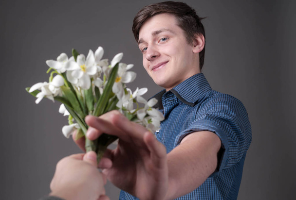 selective focus of handsome surprised and smiling young man in blue shirt taking bouquet of snowdrops from woman hand on grey background - Fotografie, Obrázek