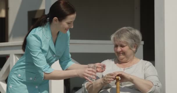 Портрет старша жінка беручи таблетки медсестра дає ліки посміхаючись разом на будинок для людей похилого віку на відкритому повітрі в сонячний день вистрілив на червону камеру - Кадри, відео