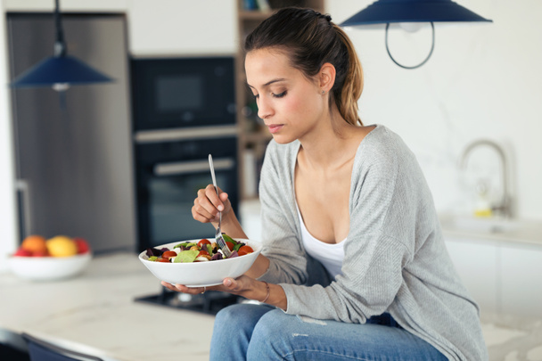 Ładna młoda kobieta jedząca sałatkę siedząc w kuchni w domu. - Zdjęcie, obraz