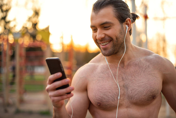 mannelijke atleet, romp met spieren, opleiding buitenshuis, lezen van SMS op de telefoon, luisteren naar muziek in koptelefoon - Foto, afbeelding