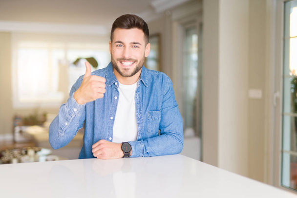 Young handsome man at home doing happy thumbs up gesture with hand. Approving expression looking at the camera with showing success. - Photo, Image