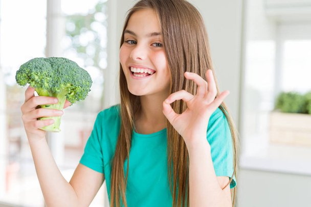 Beautiful young girl eating fresh broccoli doing ok sign with fingers, excellent symbol - Foto, Imagem