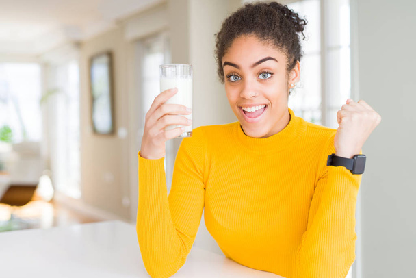 Νεαρή Αφροαμερικανή γυναίκα πίνει ένα ποτήρι φρέσκο γάλα ουρλιάζοντας περήφανη και γιορτάζει τη νίκη και την επιτυχία πολύ ενθουσιασμένη, ζητωκραυγάζοντας συγκίνηση - Φωτογραφία, εικόνα
