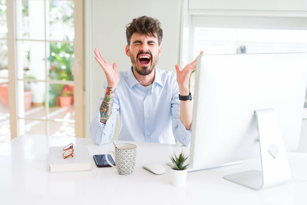 Νεαρός επιχειρηματίας που εργάζεται χρησιμοποιώντας υπολογιστή γιορτάζει τρελός και τρελός για την επιτυχία με τα χέρια ψηλά και κλειστά μάτια ουρλιάζοντας ενθουσιασμένος. Έννοια νικητή - Φωτογραφία, εικόνα