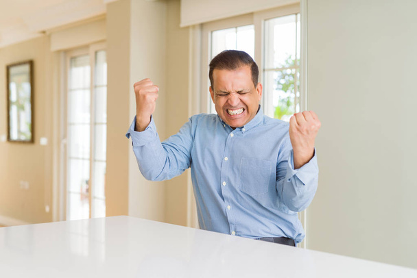 Middle age man sitting at home very happy and excited doing winner gesture with arms raised, smiling and screaming for success. Celebration concept. - Fotoğraf, Görsel