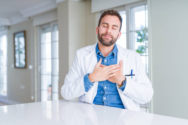 Όμορφος γιατρός άντρας φορώντας ιατρικό παλτό στην κλινική χαμογελώντας με τα χέρια στο στήθος με κλειστά μάτια και ευγνωμοσύνη χειρονομία στο πρόσωπο. Έννοια της υγείας. - Φωτογραφία, εικόνα