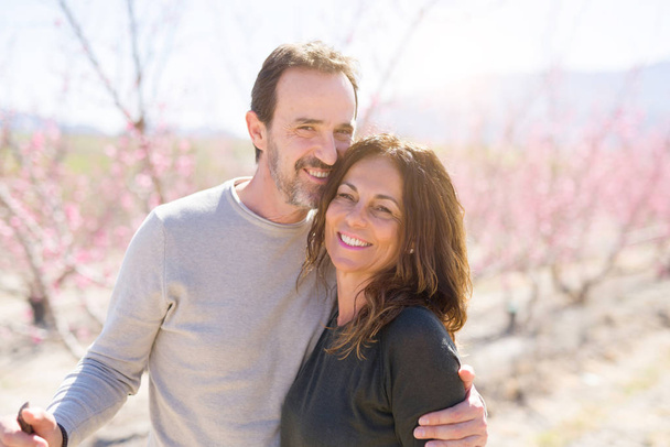 Kaunis keski-ikäinen vanhempi pari hymyilee rakkaudessa romanttisessa persikkapuiden puutarhassa vaaleanpunaisilla terälehdillä aurinkoisena kevään päivänä.
 - Valokuva, kuva