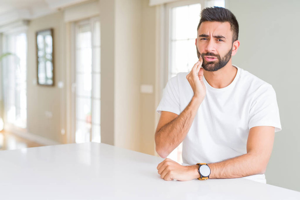 Knappe Spaanse man casual wit t-shirt thuis aanraken van de mond met de hand met pijnlijke expressie door tandpijn of tandheelkundige ziekte op tanden. Tandarts concept. - Foto, afbeelding