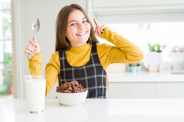 Beautiful young girl kid eating chocolate cereals and glass of milk for breakfast surprised with an idea or question pointing finger with happy face, number one - Photo, Image