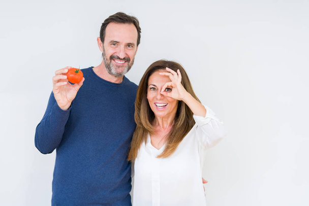 孤立した背景の上に新鮮なトマトを食べる中年のカップルは、指を通して見て目に手でOKサインをやって笑顔で笑顔 - 写真・画像