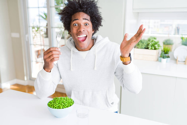 Hombre afroamericano comiendo guisantes verdes frescos en casa muy feliz y emocionado, expresión ganadora celebrando la victoria gritando con una gran sonrisa y levantando las manos
 - Foto, imagen