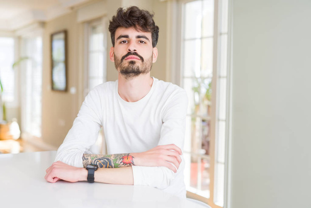 Young man wearing casual shirt sitting on white table with serious expression on face. Simple and natural looking at the camera. - Foto, Imagen