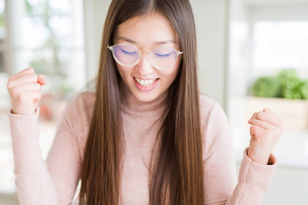 Belle femme asiatique portant des lunettes excitées pour le succès avec les bras levés célébrant la victoire en souriant. Concept gagnant
. - Photo, image