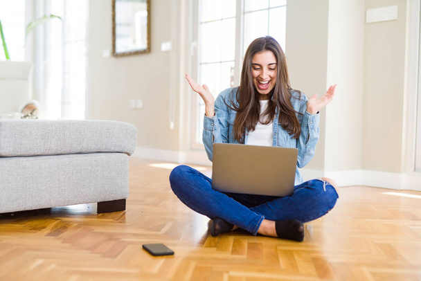 Gyönyörű fiatal nő ül a padlón keresztbe lábak segítségével laptop nagyon boldog és izgatott, győztes kifejezés ünneplő győzelmet sikoltozva nagy mosollyal és emelt kezek - Fotó, kép