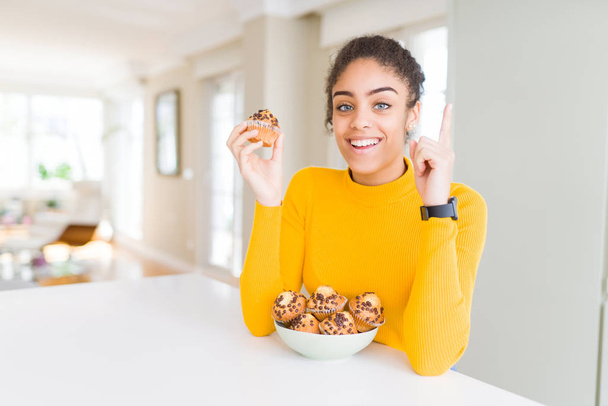Young african american woman eating chocolate chips muffins surprised with an idea or question pointing finger with happy face, number one - Foto, immagini