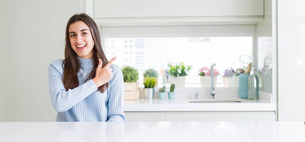 Wide angle picture of beautiful young woman sitting on white table at home cheerful with a smile of face pointing with hand and finger up to the side with happy and natural expression on face - Foto, afbeelding