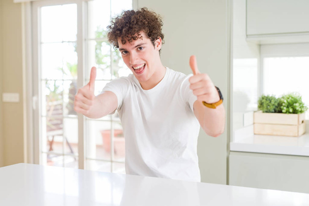 Homem bonito jovem vestindo t-shirt branca em casa aprovando fazer gesto positivo com a mão, polegares para cima sorrindo e feliz pelo sucesso. Olhando para a câmera, gesto vencedor
. - Foto, Imagem