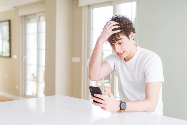Young man using smartphone at home stressed with hand on head, shocked with shame and surprise face, angry and frustrated. Fear and upset for mistake. - Photo, Image