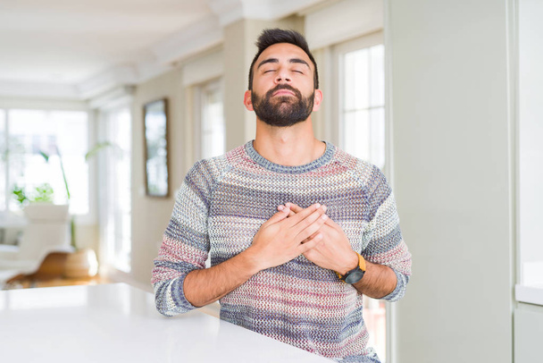 Handsome hispanic man wearing casual sweater at home smiling with hands on chest with closed eyes and grateful gesture on face. Health concept. - Fotoğraf, Görsel