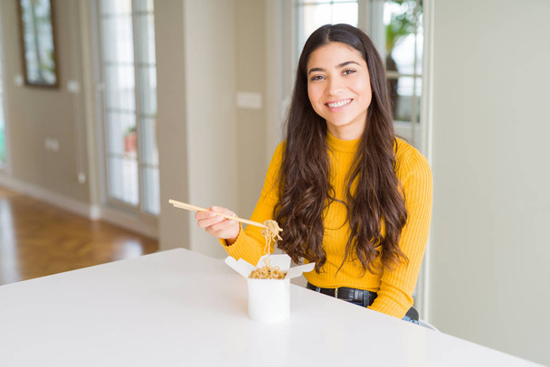 Jovem mulher comendo macarrão na caixa de entrega com um rosto feliz de pé e sorrindo com um sorriso confiante mostrando dentes
 - Foto, Imagem