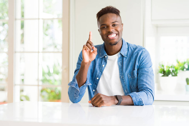 Bonito homem afro-americano em casa mostrando e apontando para cima com o dedo número um enquanto sorri confiante e feliz
. - Foto, Imagem