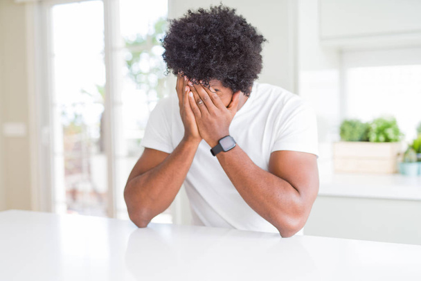 Νεαρός Αφροαμερικάνος που φοράει λευκό μπλουζάκι στο σπίτι με λυπητερή έκφραση που καλύπτει το πρόσωπο με τα χέρια ενώ κλαίει. Έννοια κατάθλιψης. - Φωτογραφία, εικόνα