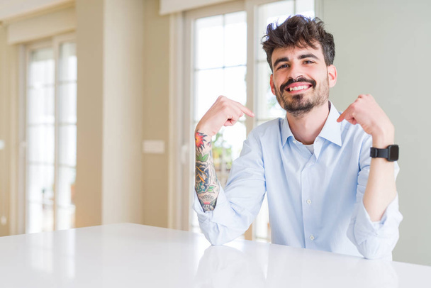 Jonge zakenman zit op witte tafel zelfverzekerd te kijken met een glimlach op zijn gezicht, wijzend met de vingers trots en gelukkig. - Foto, afbeelding