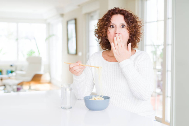 Senior woman eating asian noodles using chopsticks cover mouth with hand shocked with shame for mistake, expression of fear, scared in silence, secret concept - Foto, Bild
