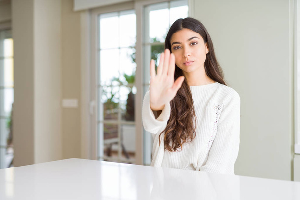 Young beautiful woman at home on white table doing stop sing with palm of the hand. Warning expression with negative and serious gesture on the face. - Photo, Image
