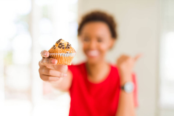 Młody african american kobieta jedzenie czekolady chipsy babeczki wskazując i Wyświetlono z kciukiem do boku z szczęśliwy twarz uśmiechający się - Zdjęcie, obraz