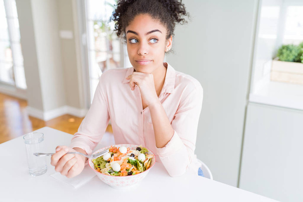 Jonge Afro-Amerikaanse vrouw die gezonde Pasta Salade eet serieus gezicht denken over vraag, zeer verward idee - Foto, afbeelding