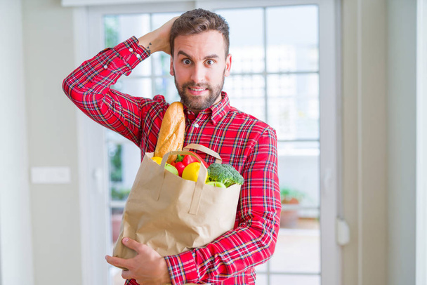 Handsome man holding groceries bag stressed with hand on head, shocked with shame and surprise face, angry and frustrated. Fear and upset for mistake. - Fotó, kép
