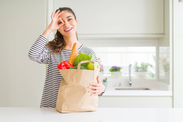 Молодая женщина держит бумажный пакет полный продуктов с счастливым лицом улыбается делает хорошо знак с рукой на глазу глядя сквозь пальцы
 - Фото, изображение