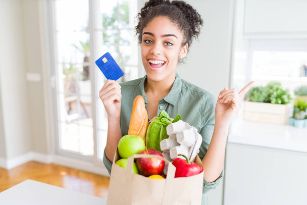 Młoda Afrykańska amerykańska dziewczyna trzyma papierową torebkę artykułów spożywczych i karty kredytowej jako płatność bardzo szczęśliwy wskazując ręką i palcem na bok - Zdjęcie, obraz